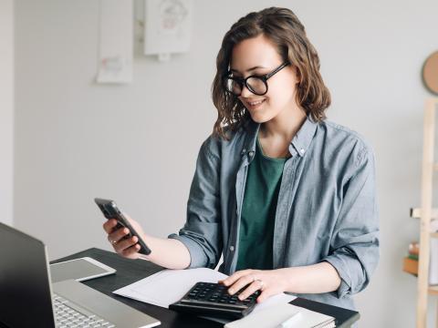 Smiling woman works at a desk using a calculator and phone, with a laptop and papers in front of her.