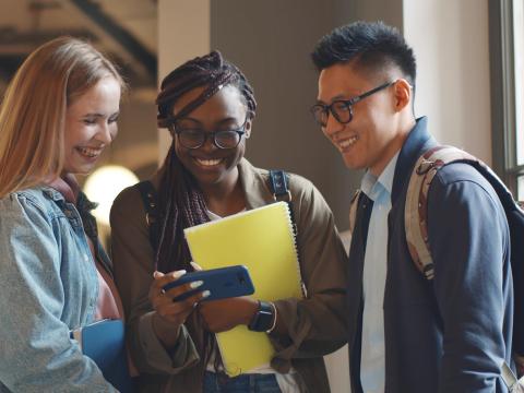 Three smiling students stand together indoors, looking at something on a phone and holding notebooks and backpacks.