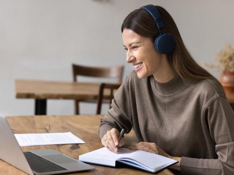 Woman wearing headphones sits at a desk, smiling while taking notes during a video call on her laptop.